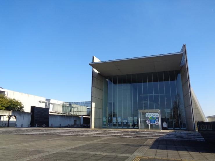 青空の下、ガラス張りの正面と張り出した屋根をもつ展示館の建物と、手前の広い石畳の広場