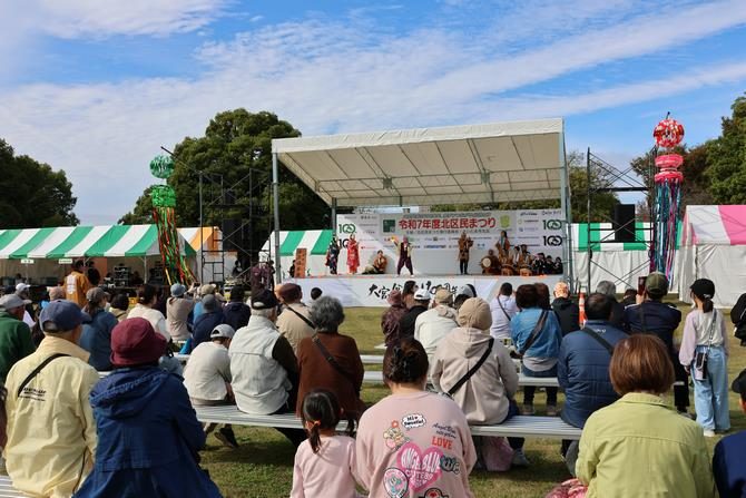 木々に囲まれた芝生広場の屋根付きステージで演奏が行われ、ベンチに座った来場者が見ている写真