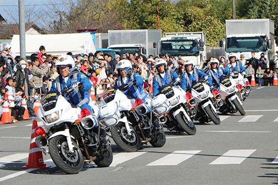 白い制服とヘルメットの隊員が乗った白バイが一列に並び、後ろに見物客が集まっている