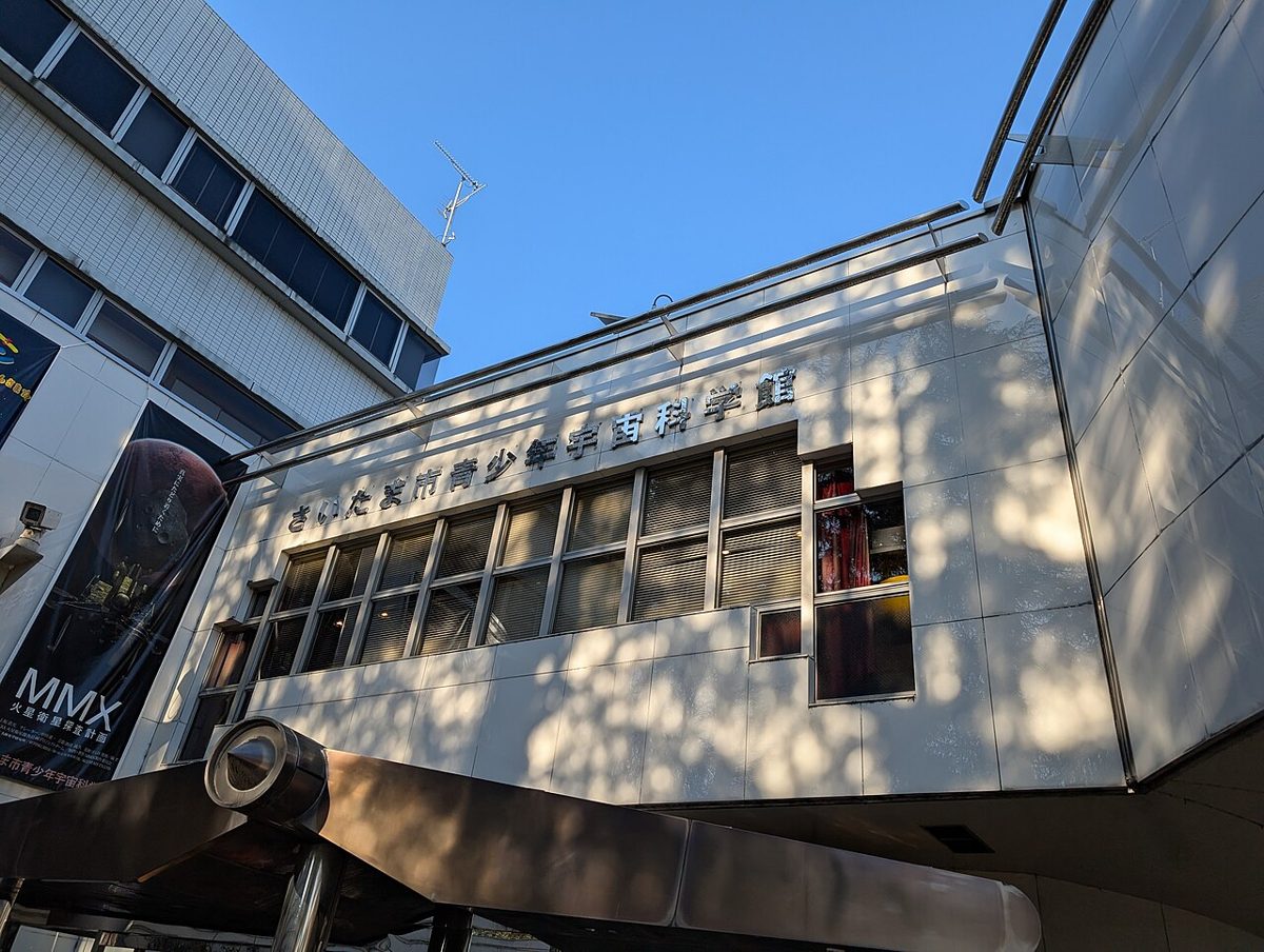 「さいたま市青少年宇宙科学館」の館名が入った白い建物の正面。左に「MMX」のプラネタリウム番組の看板