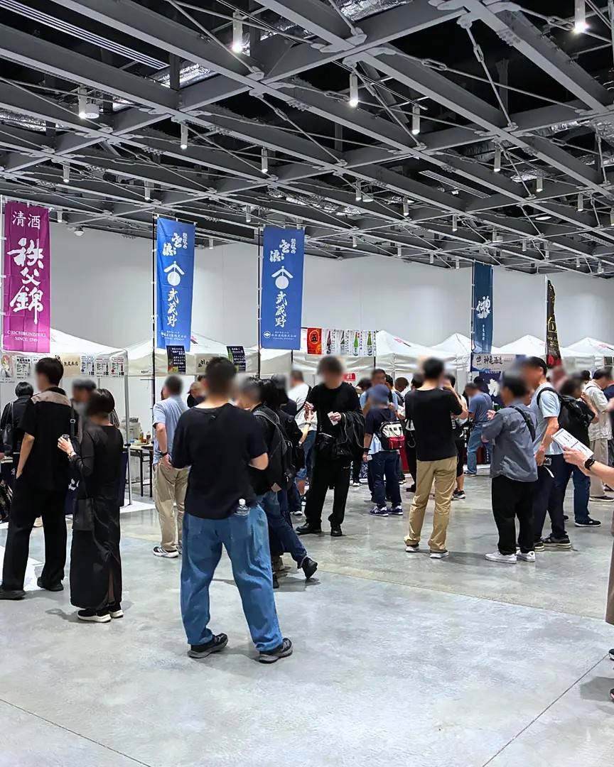 天井の高い屋内ホールに白いテントのブースが並び、秩父錦などののぼりの前で、来場者が試飲のカップを手に歩いている様子