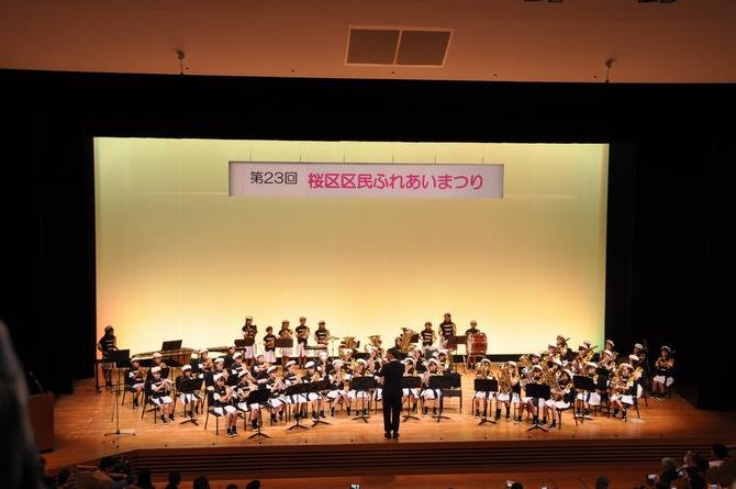 さくらホールの舞台で、小学生の金管バンドが演奏している。舞台上に「第23回桜区区民ふれあいまつり」の看板