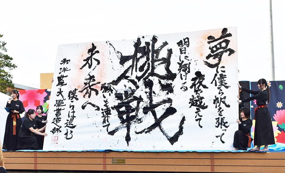 屋外ステージで黒い衣装の書道部員が、「挑戦」の大きな文字を書いた紙を立てて掲げている
