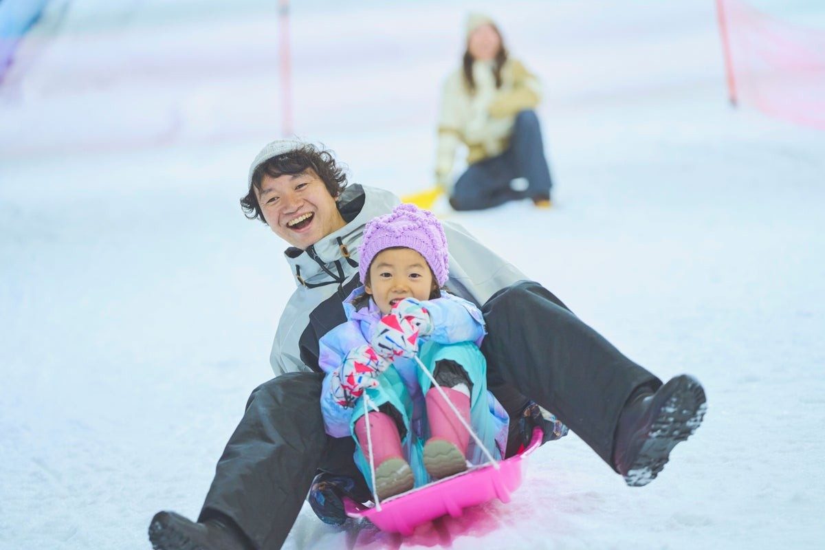 雪の斜面で、大人と帽子をかぶった子どもがピンクのそりに乗って笑顔で滑っている写真