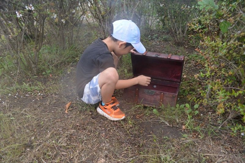 草むらに置かれた木の宝箱を、帽子をかぶった子どもがしゃがんで開けている