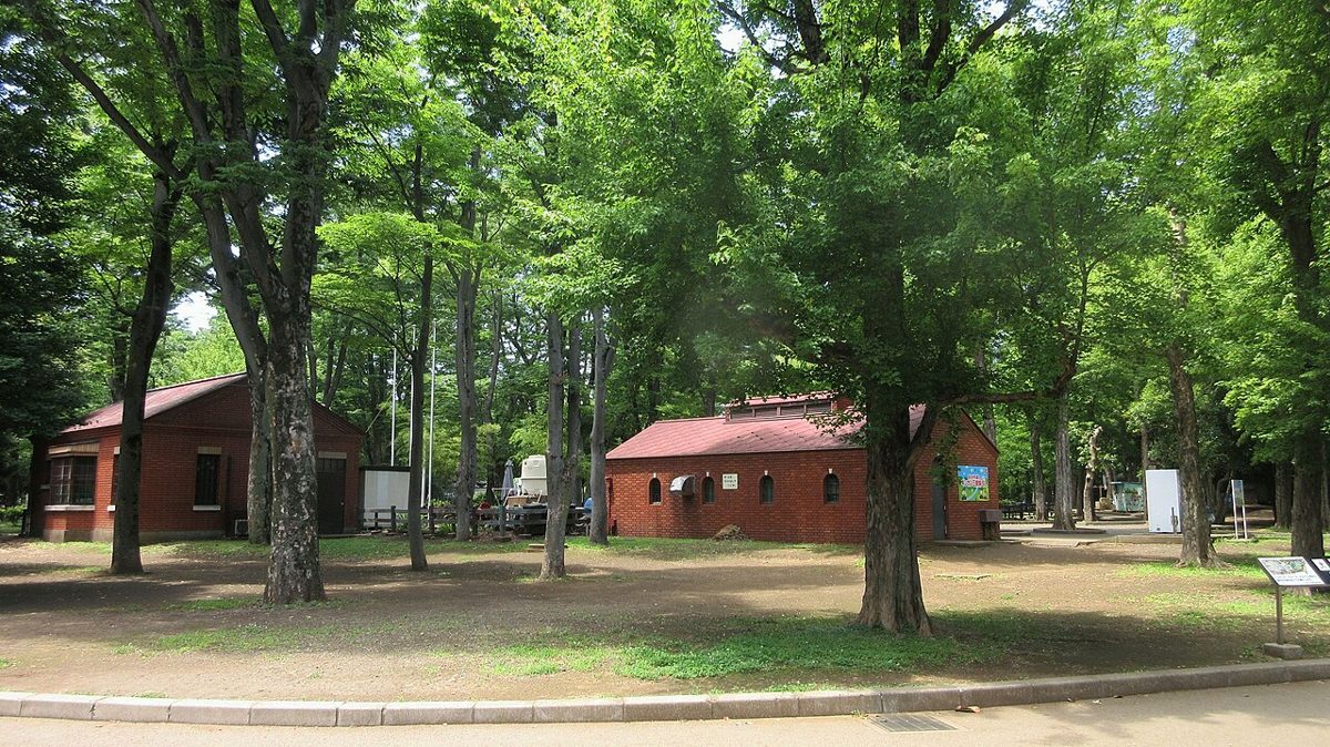 新緑の木立の中に、赤い屋根とれんが色の壁の小さな駅舎が2棟並び、手前は土の広場になっている