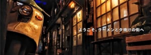 夕暮れのような照明の路地に古いスクーターと木造の店先が並び、「ようこそ、ラーメンと夕焼けの街へ」の文字