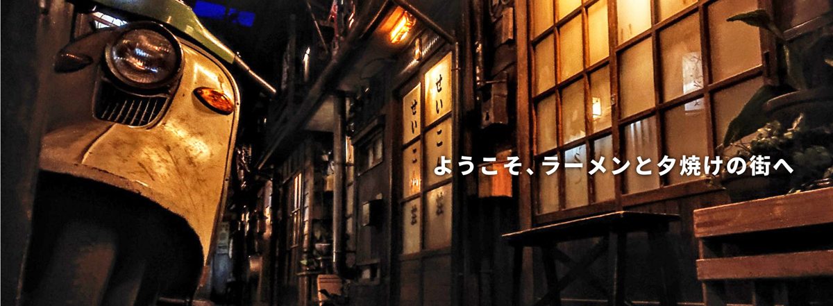 夕暮れのような照明の路地に古いスクーターと木造の店先が並び、「ようこそ、ラーメンと夕焼けの街へ」の文字