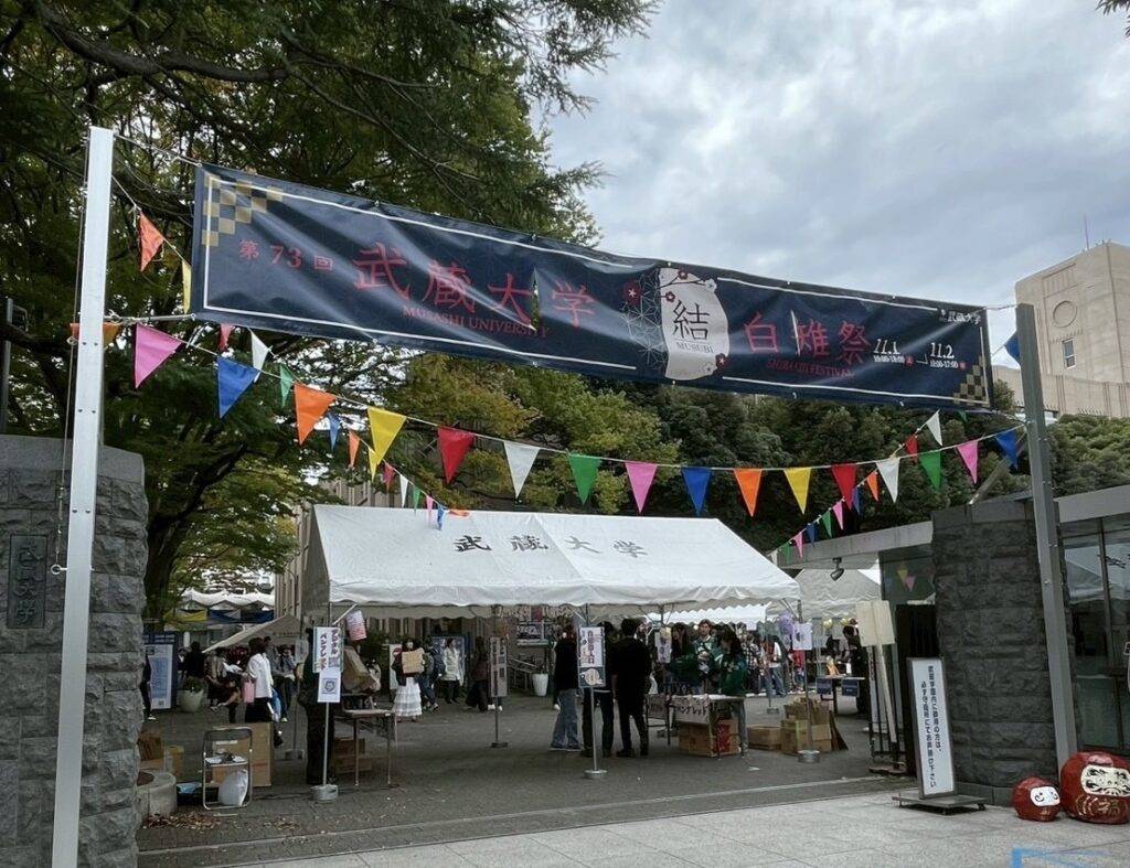 武蔵大学の正門に「第73回 武蔵大学 白雉祭 11.1 11.2」の横断幕と三角旗が飾られ、白いテントの受付に来場者が集まる様子