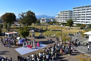 団地の建物と遠くの山を背に、広場の白い舞台でダンスを披露する人たちと、周りに集まる大勢の来場者