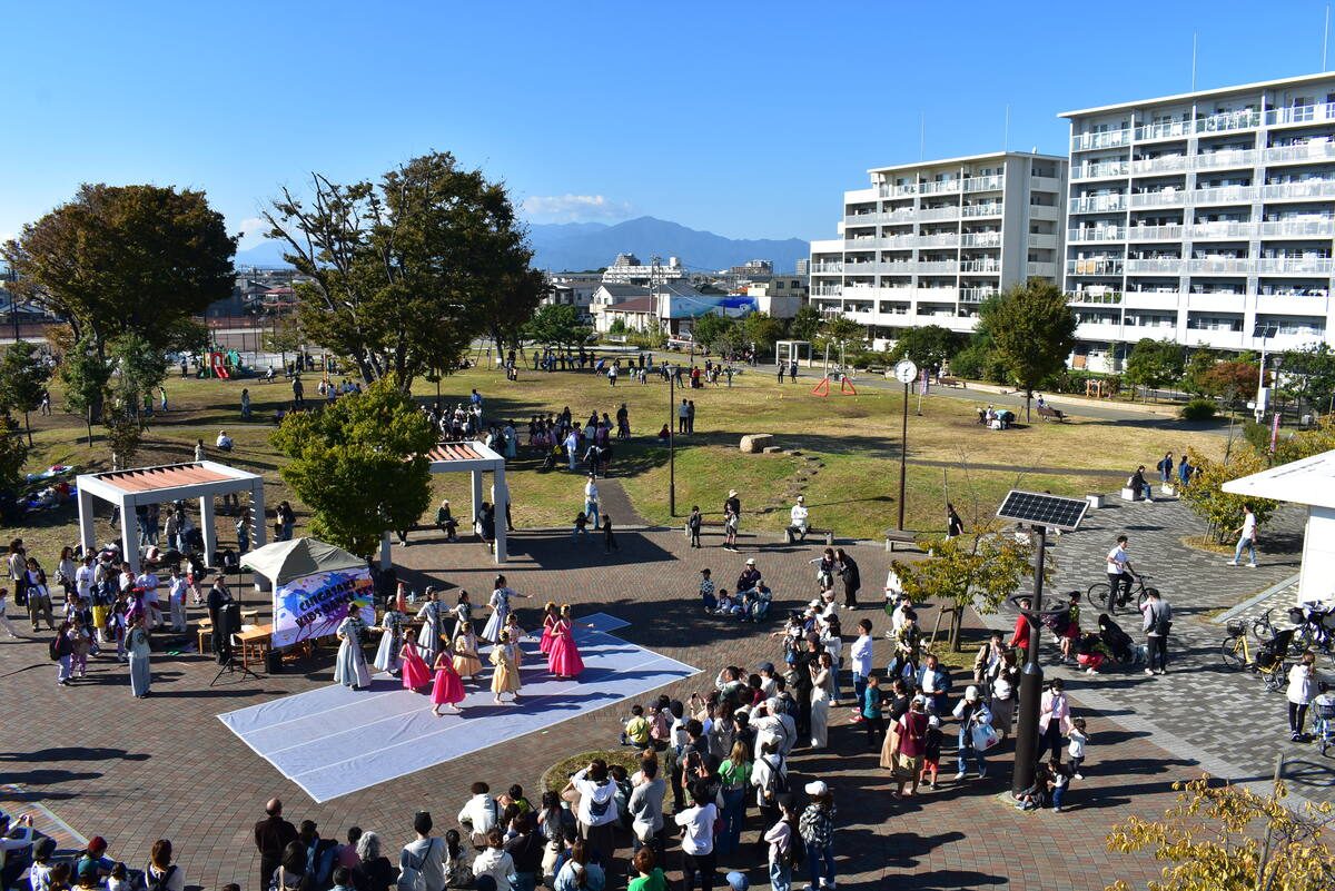 団地の建物と遠くの山を背に、広場の白い舞台でダンスを披露する人たちと、周りに集まる大勢の来場者