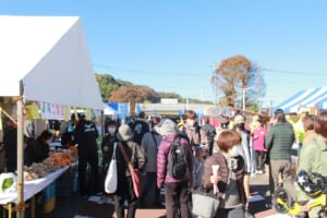 晴れた空の下、テントの露店が並ぶ通りを大勢の来場者が歩く様子。左手前のJA二宮のテントにはみかんやじゃがいもが並ぶ