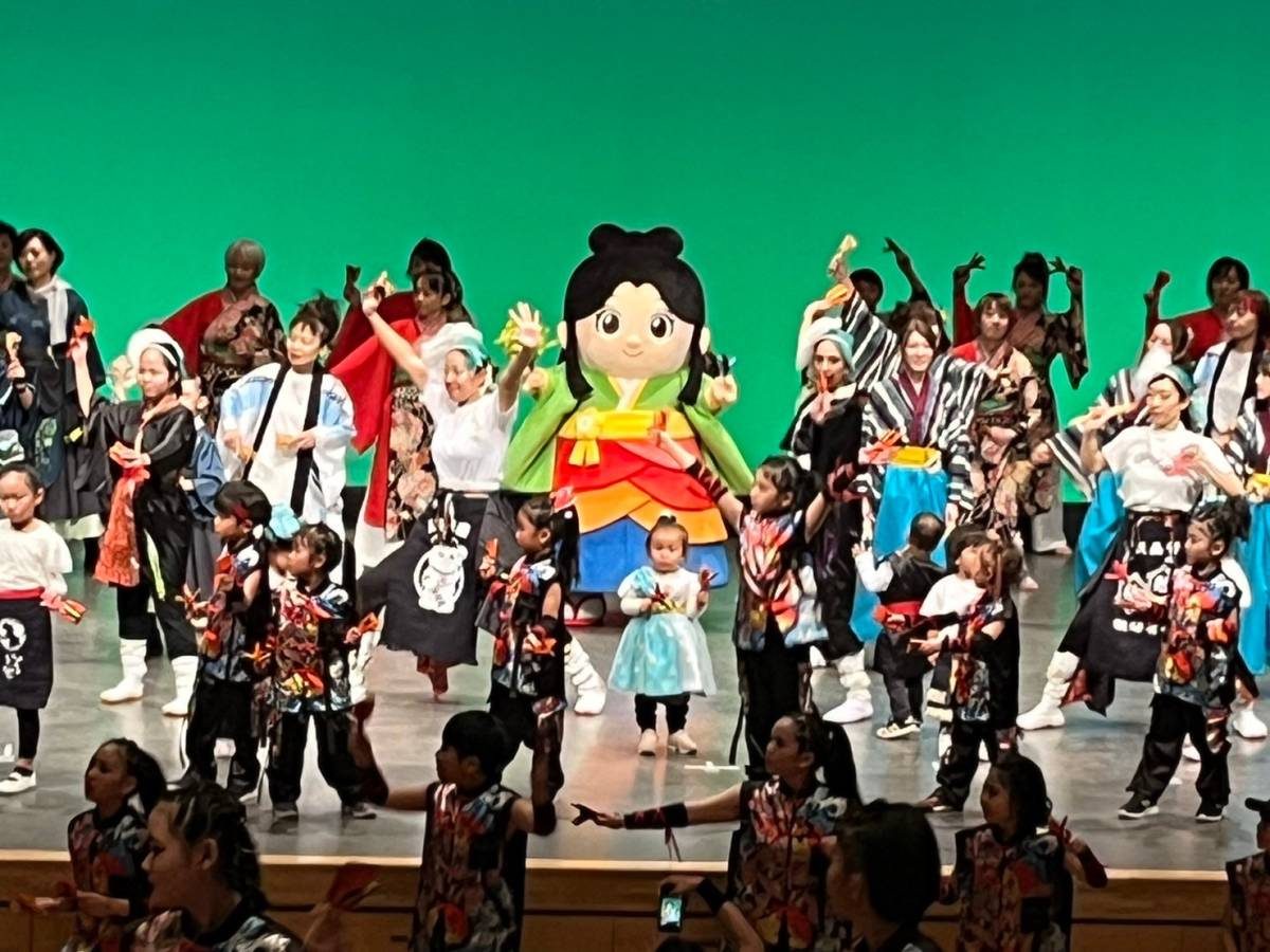 緑の幕のホールステージで、着物姿のマスコットキャラクターを囲み、法被や衣装の踊り手と子どもたちが鳴子を手に踊る様子