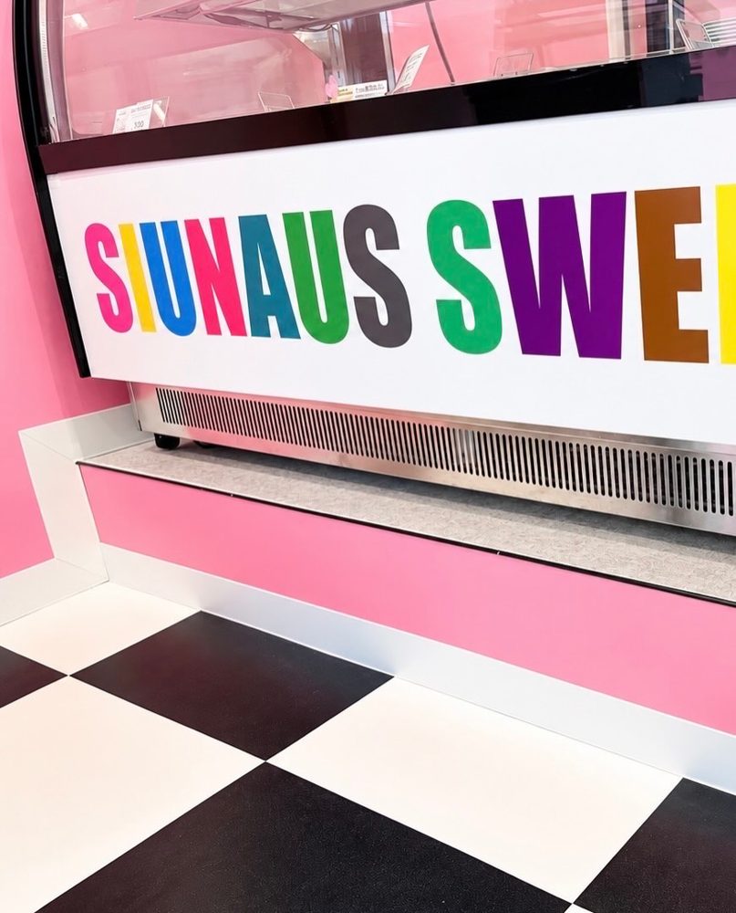 ピンクの壁と白黒の市松模様の床の前に、カラフルな「SIUNAUS SWEETS」のロゴが入ったショーケース