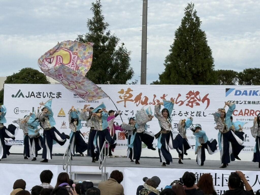 草加ふささら祭りのメインステージで、よさこいの踊り手が大きな旗を振って演舞し、手前で観客が見ている