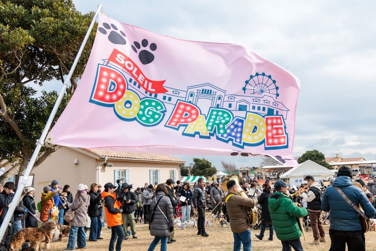 SOLEIL DOG PARADEの旗の下に犬を連れた参加者が集まる様子