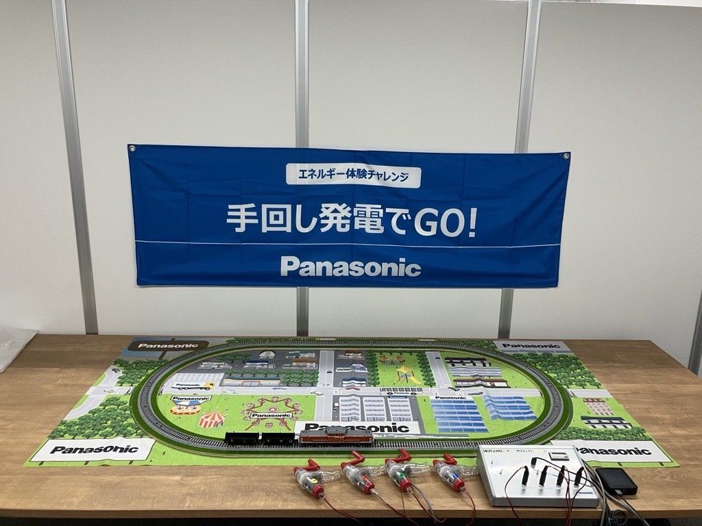 「エネルギー体験チャレンジ 手回し発電でGO!」の幕の下、テーブルに置かれた街のジオラマと線路、電車の模型、手回し発電機