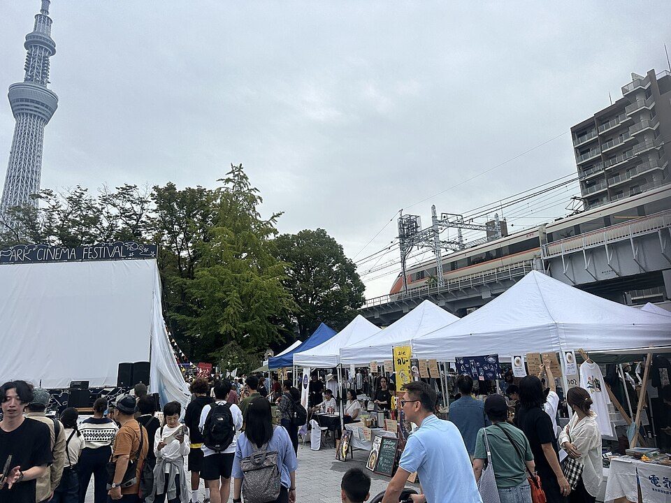 曇り空の下、左奥にスカイツリー、右に東武線の高架と白いテントが並び、来場者が行き交う隅田公園の広場