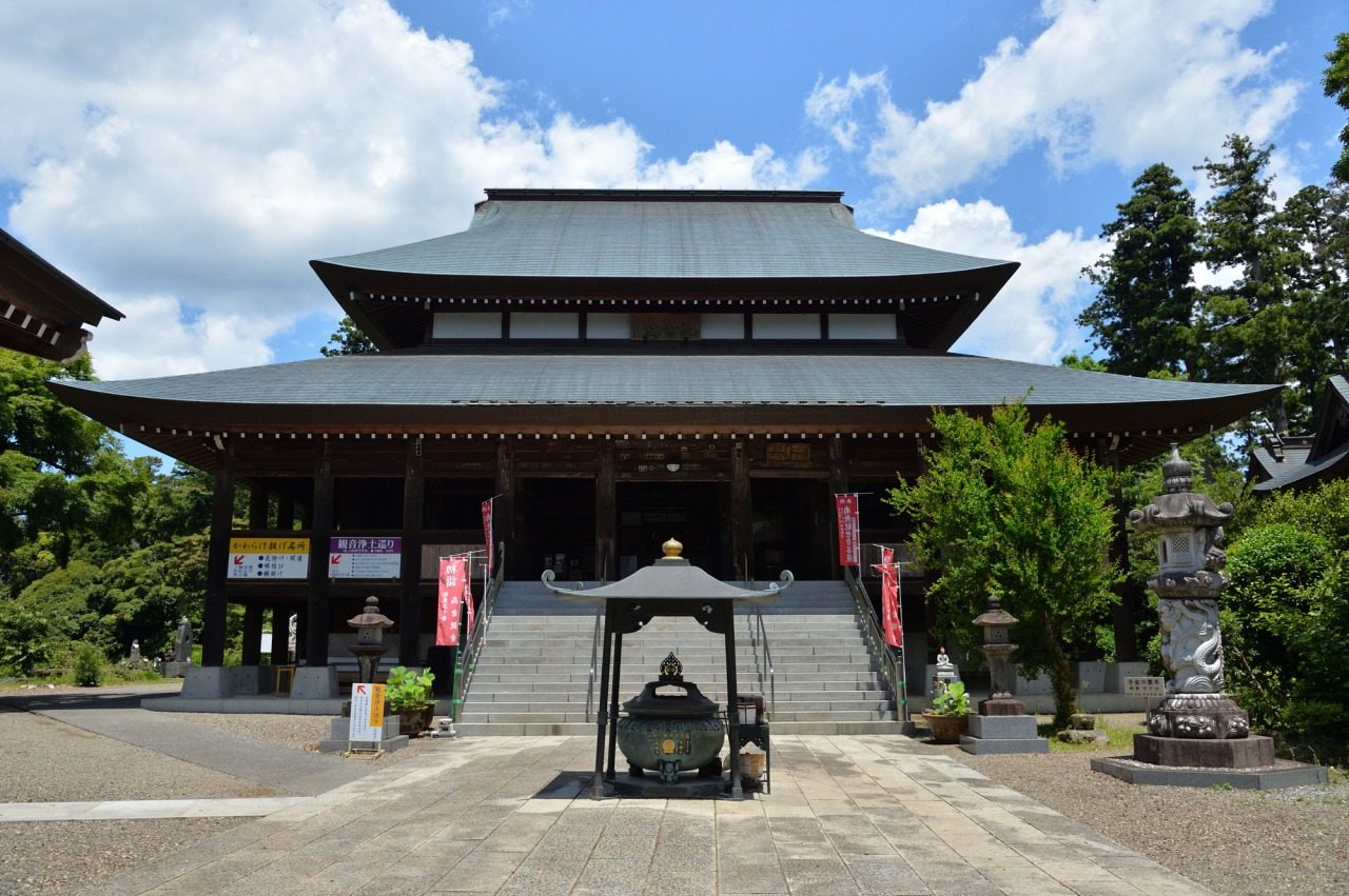 青空の下の高蔵寺の本堂。二重の屋根の大きな木造のお堂に石段が続き、手前に香炉、両脇に石灯籠と赤いのぼり旗