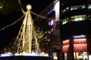 光のカーテンを重ねた円すい形のツリーと、ツリーの頂点から左右へ延びる電飾。右奥にココリア多摩センターの建物