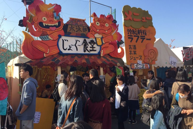 鬼の顔の大きな看板と手描きのメニューで飾った模擬店「じょっぐ食堂」の前に、来場者が集まっている様子
