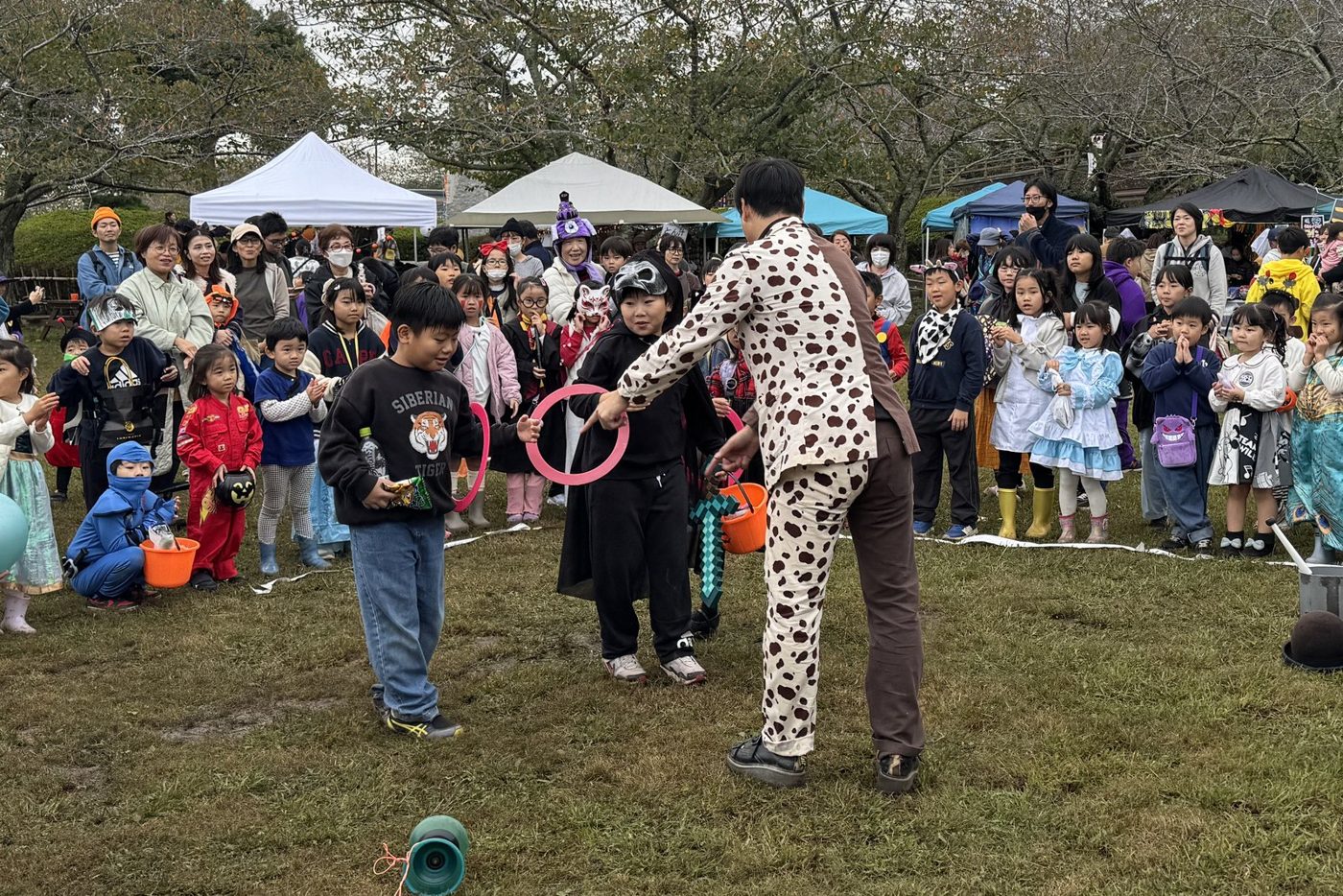 2025年の館山ハロウィンフェスで、城山公園の芝生広場に仮装した子どもたちが集まり、水玉模様の衣装の大道芸人ケンケンさんが子どもに輪を持たせている様子。後ろに出店のテントが並んでいる