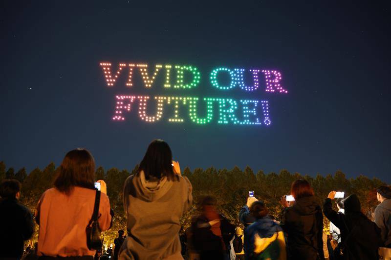 夜空にドローンの光で描かれた「VIVID OUR FUTURE!」の虹色の文字と、それをスマートフォンで撮影する来場者の後ろ姿