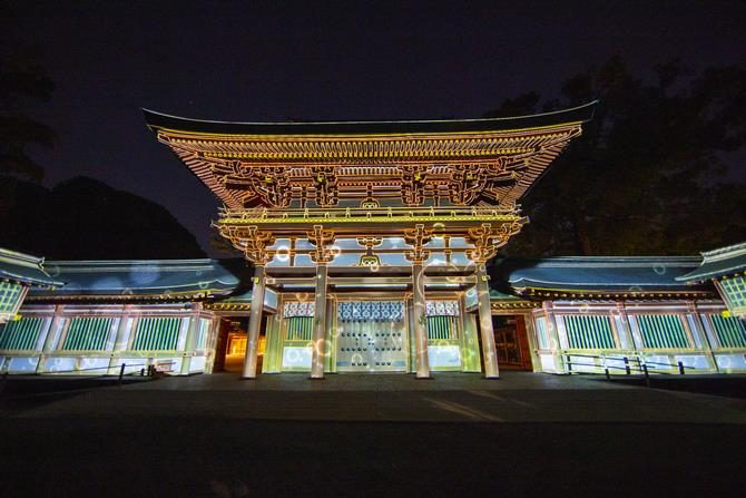 夜の暗がりの中で、朱色の楼門と左右の回廊が白い光で照らされている写真