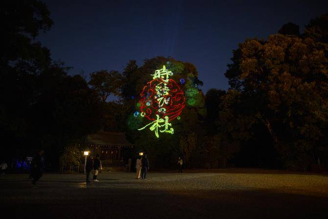 夜の境内の木に、赤い円の模様と「時結びの杜」の文字が緑色で投影され、手前を人が歩いている写真