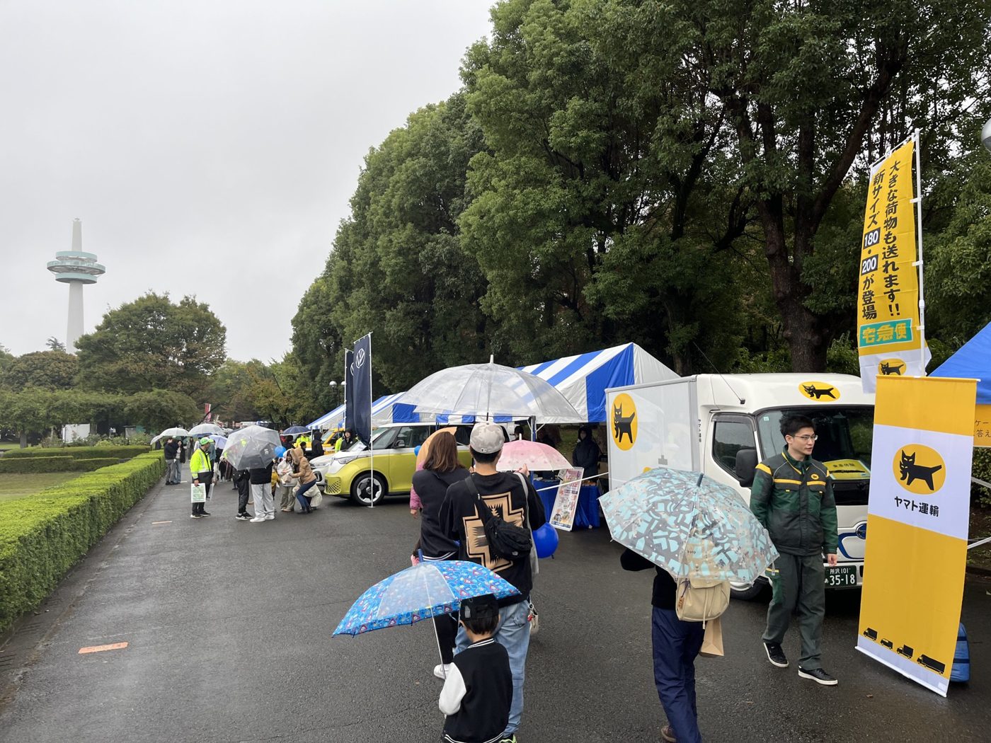雨の中、傘を差して出展テントの並ぶ通路を歩く来場者。奥に航空公園の放送塔