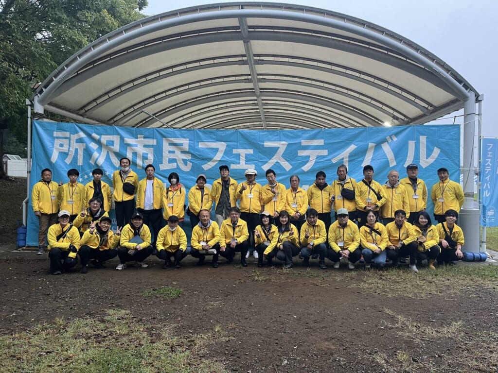 曇り空の下、「所沢市民フェスティバル」の横断幕を掲げたステージ前に並ぶ黄色いジャンパーの実行委員たち