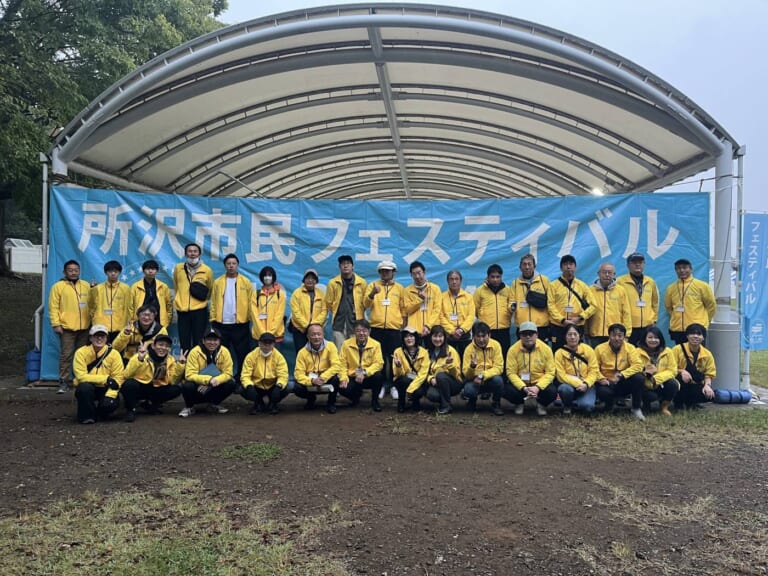 曇り空の下、「所沢市民フェスティバル」の横断幕を掲げたステージ前に並ぶ黄色いジャンパーの実行委員たち