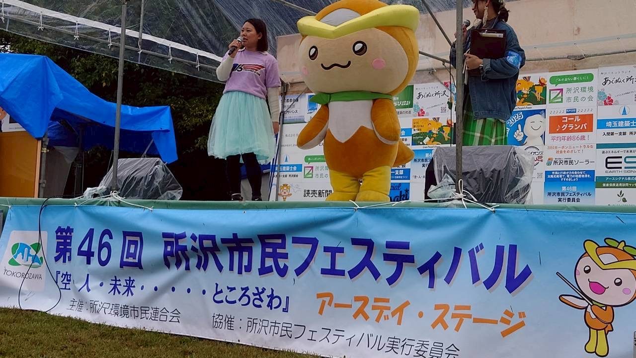 「第46回所沢市民フェスティバル」の幕がかかったテントのステージで、歌う出演者とゆるキャラのトコろん