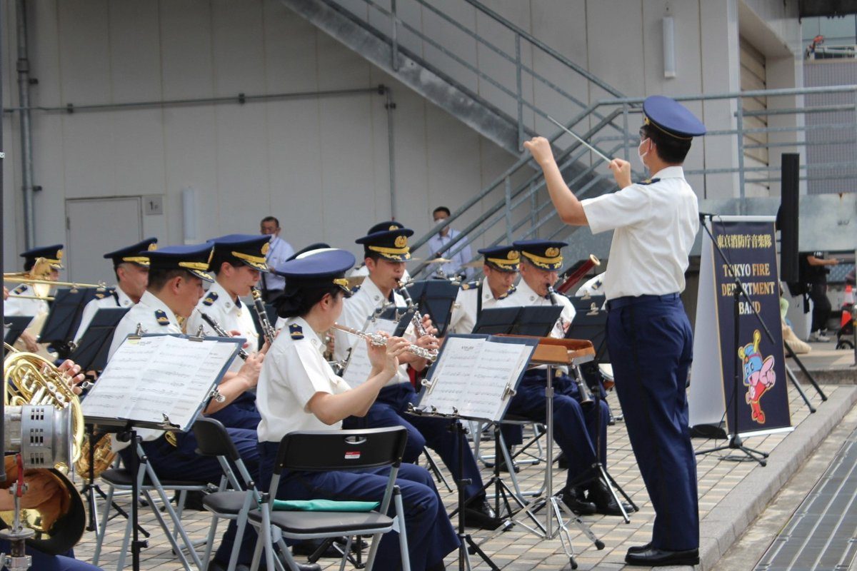制服姿の東京消防庁音楽隊が屋外で演奏し、指揮者がタクトを振っている