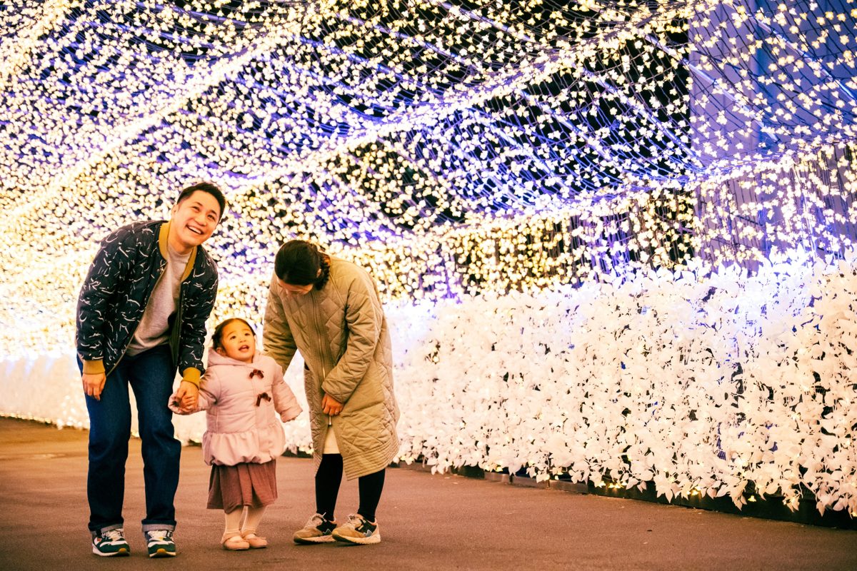 白い電球に覆われた光のトンネルの中で、男性と女性が小さな女の子と手をつないで歩いている
