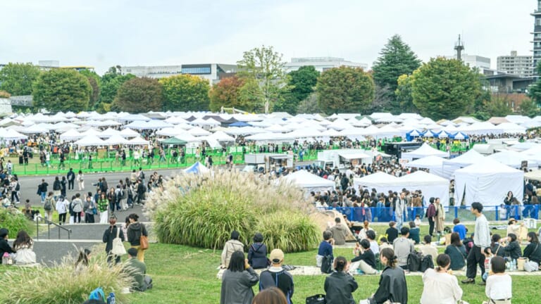 昭和記念公園のゆめひろばに白いテントが一面に並び、多くの来場者が歩き、手前の芝生の丘に座って休む人たちがいる東京蚤の市の会場の写真