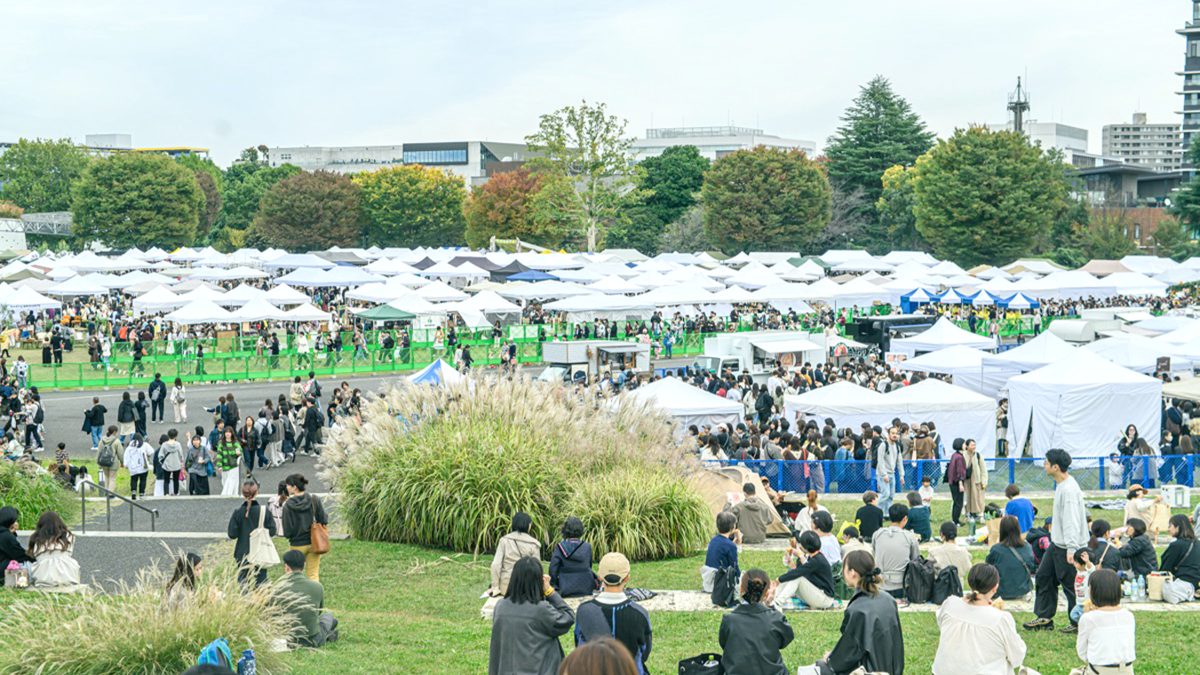 昭和記念公園のゆめひろばに白いテントが一面に並び、多くの来場者が歩き、手前の芝生の丘に座って休む人たちがいる東京蚤の市の会場の写真