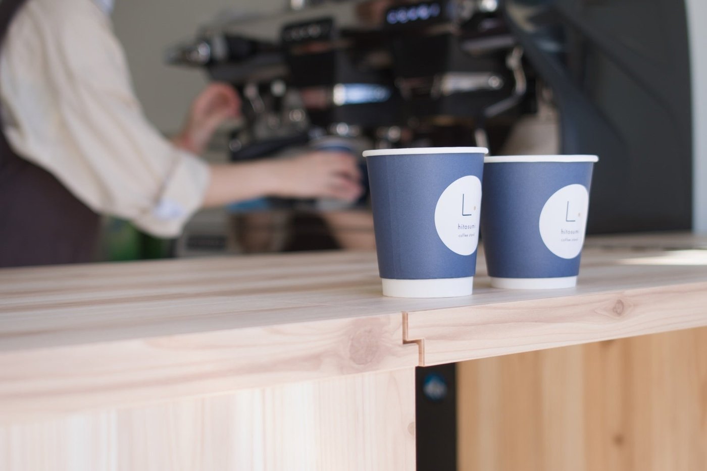 木目のカウンターに青い紙カップが2個並び、奥でスタッフが作業する店内