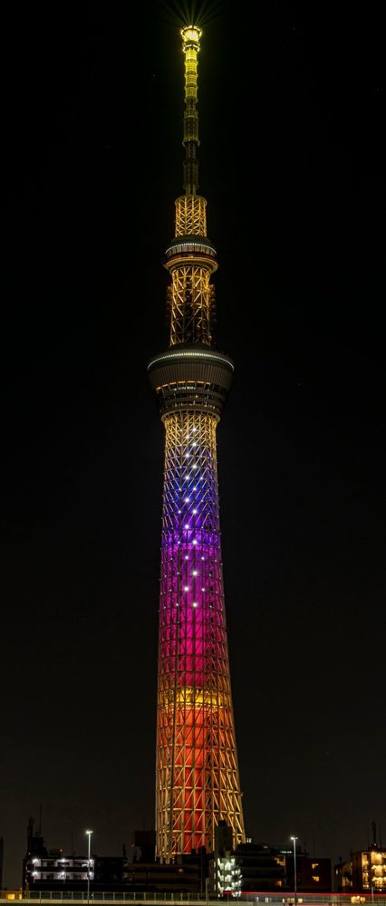 夜空の下で、塔の上部が淡い黄色、中ほどから下が紫やオレンジのグラデーションに光る東京スカイツリー