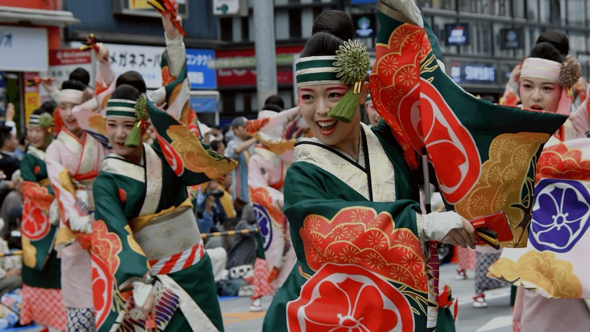 池袋駅西口の通りで、緑と白の衣装に赤い花柄の着物を重ねた踊り子たちが鳴子を手に踊る様子
