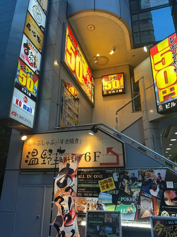 夕方のビルの入口。外壁や入口上に黄色地に赤字で「レモンサワー50円」と書いた看板が並び、階段が上へ続いている写真