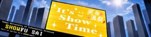 高層ビル街を背景にした看板に「It's Show Time」「笑顔満祭」、下に「SHOWFU SAI」の文字
