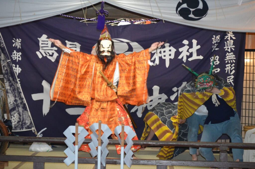鳥見神社の神楽殿で舞う浦部の神楽。「浦部神楽 十二座」「鳥見神社」と染め抜いた幕の前で、橙の装束の舞手が両腕を広げ、右手に獅子頭のような面の舞手が控える