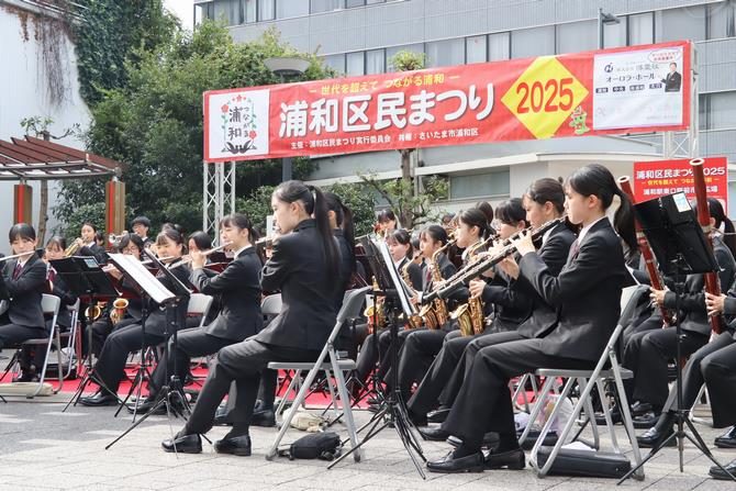 「浦和区民まつり2025」の横断幕の下で、制服姿の高校生が吹奏楽を演奏している浦和駅東口のステージの写真