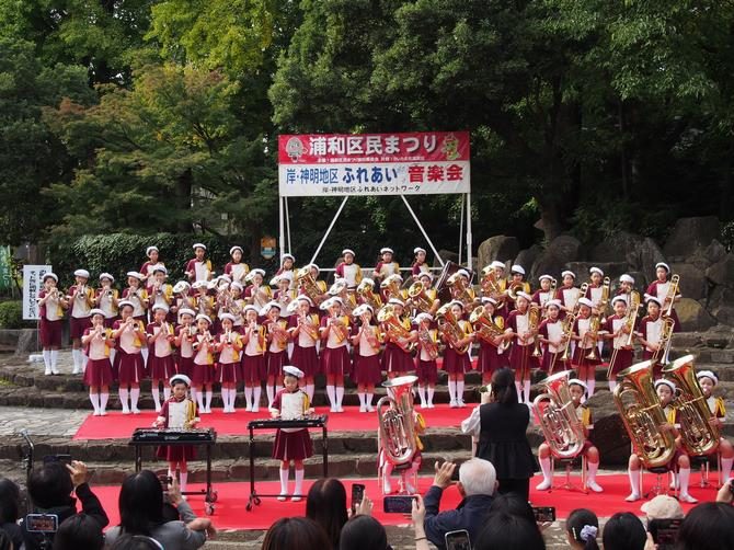 「浦和区民まつり 岸・仲町地区 ふれあい音楽会」の横断幕の前で、赤い制服の子どもたちが金管楽器を演奏している写真