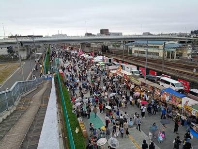 浦和美園駅横の歩行者専用道路に屋台のテントが並び、大勢の来場者が行き交っている。奥に線路と高架