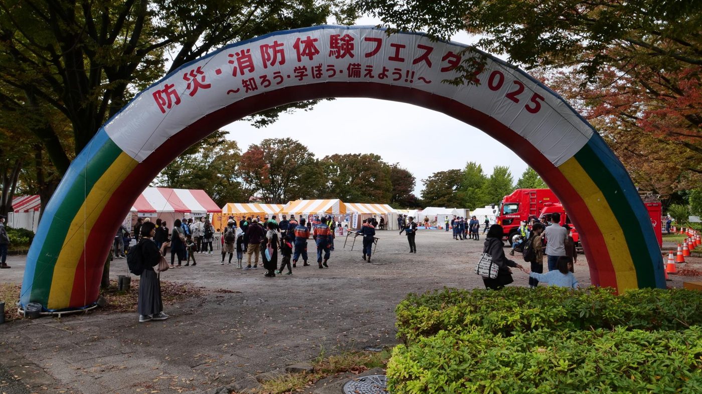 「防災・消防体験フェスタ2025」と書かれた虹色のアーチと、その奥に並ぶテントや消防車、来場者
