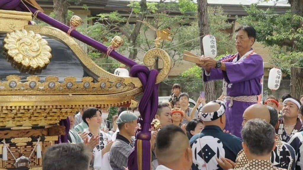 2023年の八幡祭。葛飾八幡宮の境内で、金色の装飾と紫の綱の宮神輿の前に法被姿の担ぎ手が集まり、紫の装束の男性が拍子木を打っている