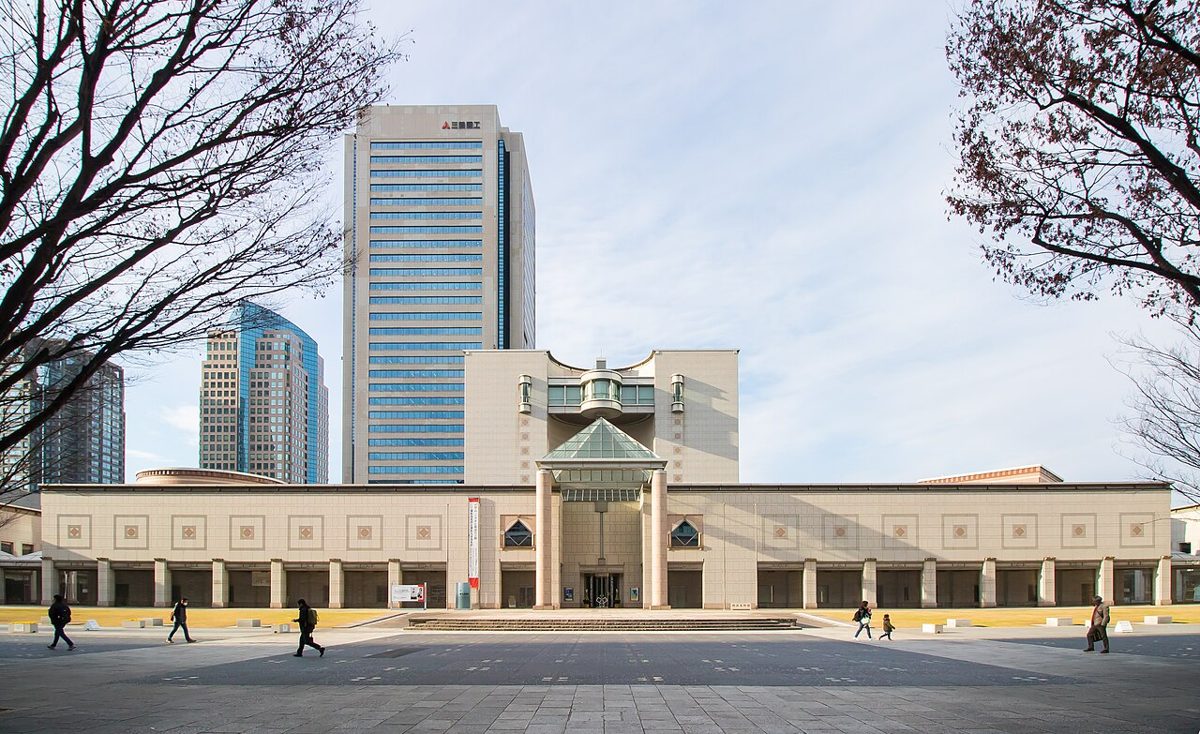 広場の奥に横長のベージュ色の建物と中央の塔が立ち、背後にみなとみらいの高層ビルが見える