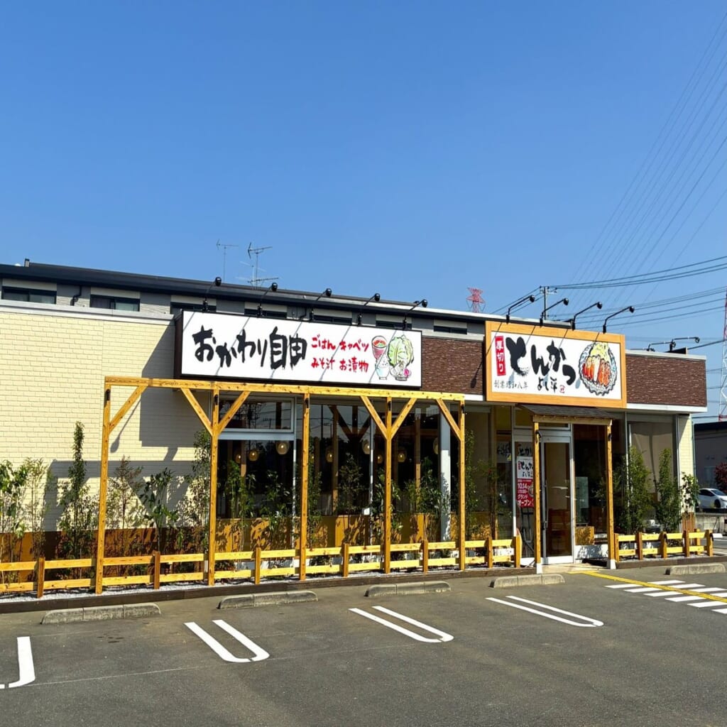 黄色い外壁の平屋の店舗。「おかわり自由」の看板と「とんかつ よし平」の看板が掲げられ、手前に白線を引いた駐車場がある